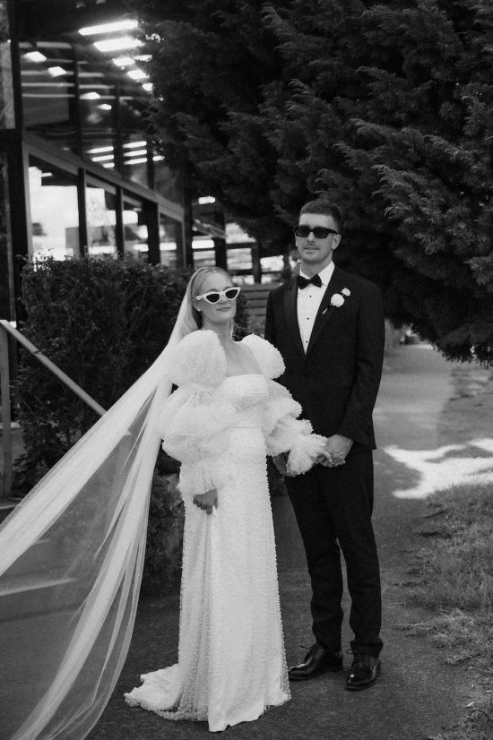 Bride in statement Cristie Nicole wedding dress covered in pearls standing with the groom dressed in a black tuxedo at The Glasshouse Morningside Auckland City Wedding Ana Galloway