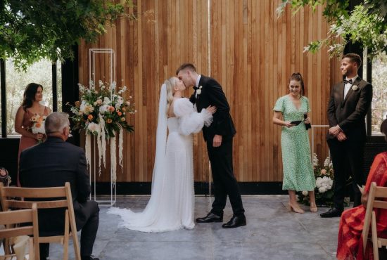Wedding ceremony first kiss bride wears a statement Cristie Nicole wedding dress covered in pearls standing with the groom dressed in a black tuxedo at The Glasshouse Morningside Auckland City Wedding Ana Galloway