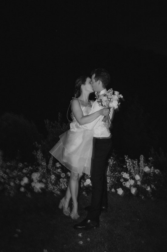 Photographic flash on the couple as the elegant bride and groom embrace and kiss at night in front of the ceremony flowers Hawkes Bay Wedding Ana Galloway Photography