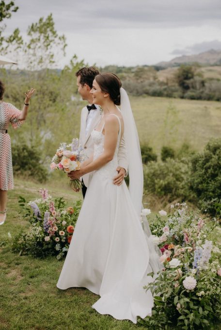 Elegant bride and groom stand in front of the wedding ceremony flowers with the picturesque backdrop of Te Mata Peak in Hawkes Bay Ana Galloway Photography