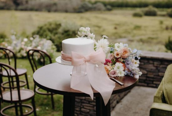 Single tier wedding cake iced with white icing and wrapped in a large blush chiffon ribbon surrounded by colourful wild flowers Hawkes Bay Wedding Ana Galloway Photography