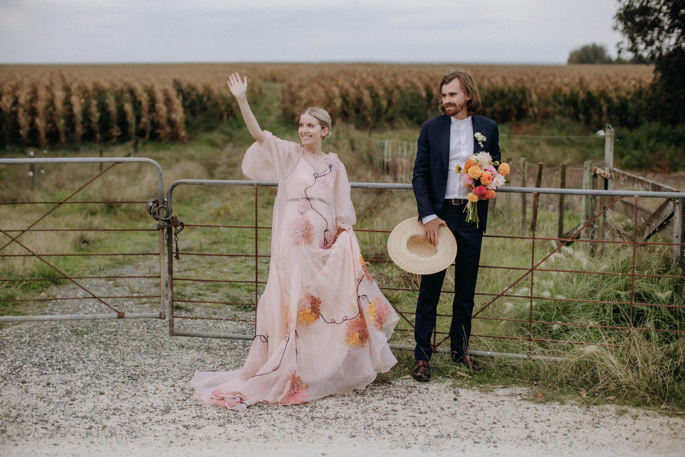 Bride wears couture blush wedding gown and groom stands next to her at a farm gate as they wave at someone. Hawkes Bay Wedding Ana Galloway Photography