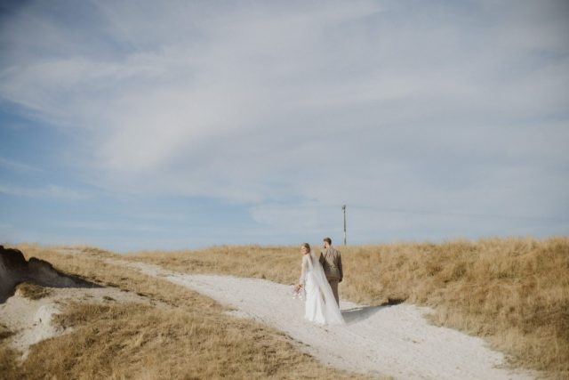 Bride and groom walking along a white dust road amongst the golden summery grass in Taupo Wedding Ana Galloway Photography