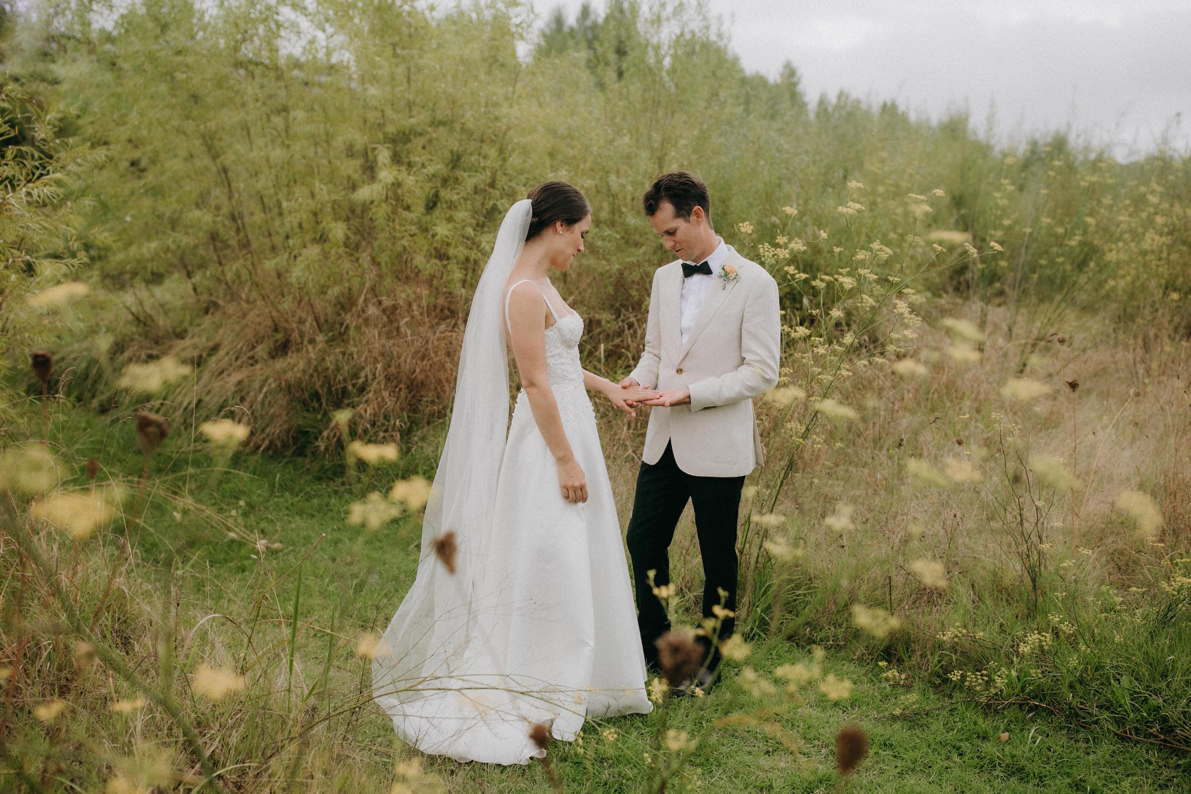 Elegant bride and groom softly hold hands amongst soft green grasses and yellow flowers Hawkes Bay Wedding Ana Galloway Photography