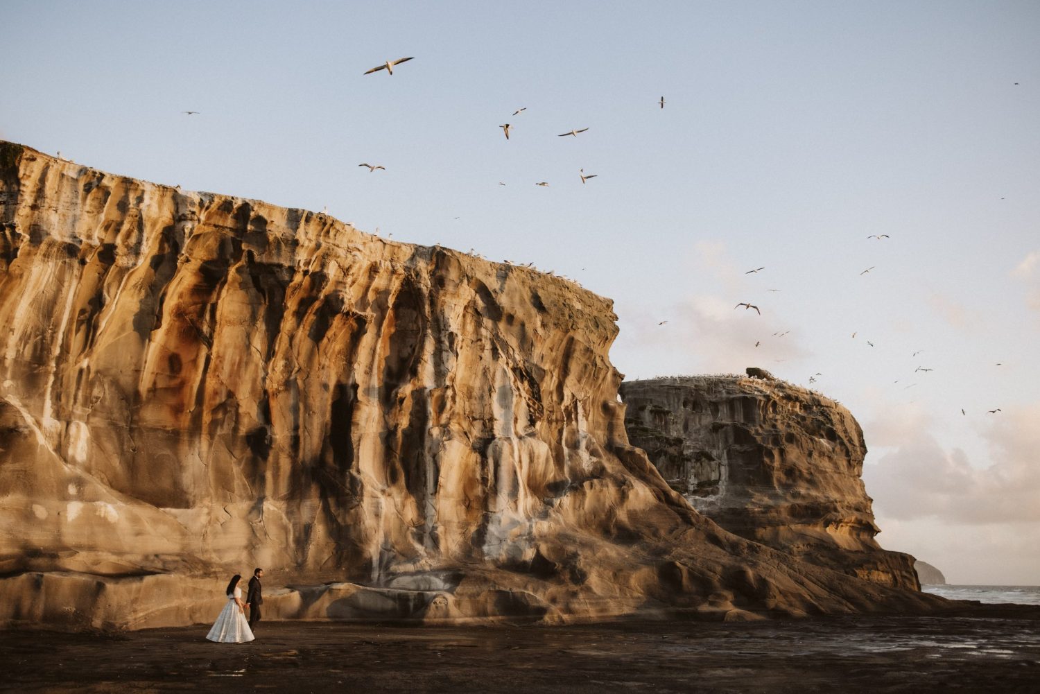 Birds flying above groom and bride wearing a modern Indian inspired dress as they walk beneath the impressive cliffs of Muriwai Beach Auckland Ana Galloway Photography